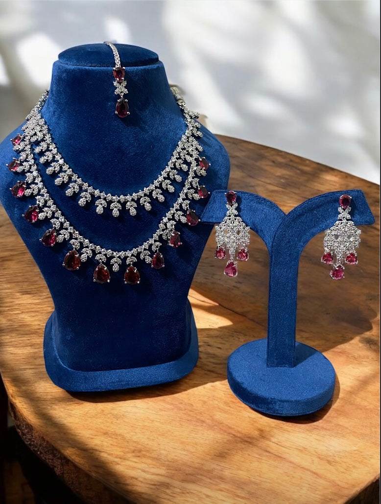 A designer American diamond necklace with double layers and vibrant ruby red gemstones displayed on a blue velvet stand, accompanied by matching earrings.